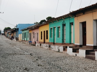 A row of colorful, single-story houses lines a cobblestone street. Each house is painted in bright colors such as blue, green, yellow, and peach. The street is quiet with a few people in the distance.
