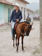 A man wearing a white cowboy hat and blue shirt rides a brown horse on a dirt road in a rural town. He is holding a can in one hand. Several other people on horseback are visible in the background, and colorful buildings line the street.
