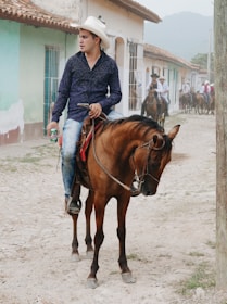 A man wearing a white cowboy hat and blue shirt rides a brown horse on a dirt road in a rural town. He is holding a can in one hand. Several other people on horseback are visible in the background, and colorful buildings line the street.