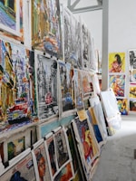A collection of vibrant and colorful paintings displayed on a makeshift wall. The artworks feature a variety of subjects, including vintage cars, urban street scenes, and portraits. Some pieces use bold colors while others are in monochrome. The setting appears to be an art studio or gallery space with a high ceiling and industrial elements.