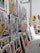 A collection of vibrant and colorful paintings displayed on a makeshift wall. The artworks feature a variety of subjects, including vintage cars, urban street scenes, and portraits. Some pieces use bold colors while others are in monochrome. The setting appears to be an art studio or gallery space with a high ceiling and industrial elements.