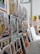 A collection of vibrant and colorful paintings displayed on a makeshift wall. The artworks feature a variety of subjects, including vintage cars, urban street scenes, and portraits. Some pieces use bold colors while others are in monochrome. The setting appears to be an art studio or gallery space with a high ceiling and industrial elements.