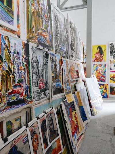 A collection of vibrant and colorful paintings displayed on a makeshift wall. The artworks feature a variety of subjects, including vintage cars, urban street scenes, and portraits. Some pieces use bold colors while others are in monochrome. The setting appears to be an art studio or gallery space with a high ceiling and industrial elements.