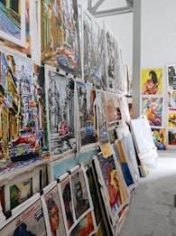 A collection of vibrant and colorful paintings displayed on a makeshift wall. The artworks feature a variety of subjects, including vintage cars, urban street scenes, and portraits. Some pieces use bold colors while others are in monochrome. The setting appears to be an art studio or gallery space with a high ceiling and industrial elements.