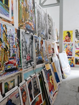 A collection of vibrant and colorful paintings displayed on a makeshift wall. The artworks feature a variety of subjects, including vintage cars, urban street scenes, and portraits. Some pieces use bold colors while others are in monochrome. The setting appears to be an art studio or gallery space with a high ceiling and industrial elements.
