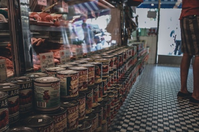 A row of canned tomatoes is neatly stacked on a tiled floor near a butcher's counter displaying sausages and meat. The scene appears to be inside a delicatessen or grocery store with soft lighting and a vintage aesthetic.