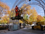 A worker wearing bright orange safety gear is operating a yellow cherry picker lift under a metal structure in an outdoor setting. The scene is surrounded by trees with vibrant autumn foliage, and there are several bicycles parked nearby.