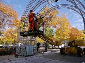 A worker wearing bright orange safety gear is operating a yellow cherry picker lift under a metal structure in an outdoor setting. The scene is surrounded by trees with vibrant autumn foliage, and there are several bicycles parked nearby.