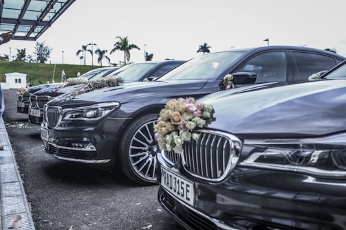 A row of luxury cars parked in an outdoor setting, each adorned with floral arrangements on their fronts. The vehicles are lined up under a modern canopy with a background of palm trees, suggesting a formal or celebratory event.