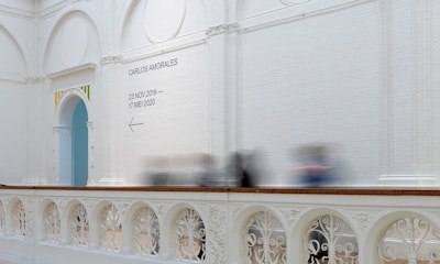 A spacious gallery with high white brick walls features a decorative archway trimmed with colorful accents. A sign on the wall provides information about an exhibition by Carlos Amorales, scheduled from 23 November 2019 to 17 May 2020. Below the sign, an arrow points to the left. The balustrade exhibits elaborate ornamental patterns. Several blurred figures indicate the presence of moving visitors.