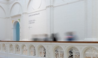 A spacious gallery with high white brick walls features a decorative archway trimmed with colorful accents. A sign on the wall provides information about an exhibition by Carlos Amorales, scheduled from 23 November 2019 to 17 May 2020. Below the sign, an arrow points to the left. The balustrade exhibits elaborate ornamental patterns. Several blurred figures indicate the presence of moving visitors.