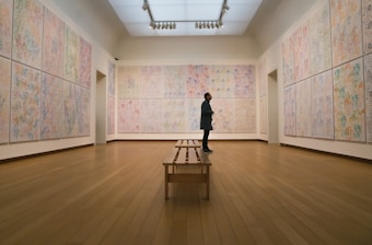 A person stands in a spacious art gallery, surrounded by walls adorned with large, colorful abstract paintings. The floor is wooden and polished, leading to a bench centered in the room. The lighting is soft and evenly distributed, highlighting the artwork.