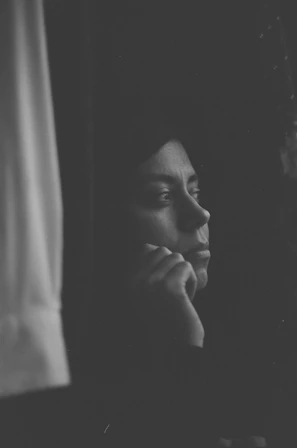 Black and white photo of a thoughtful author gazing out a window, pen in hand.