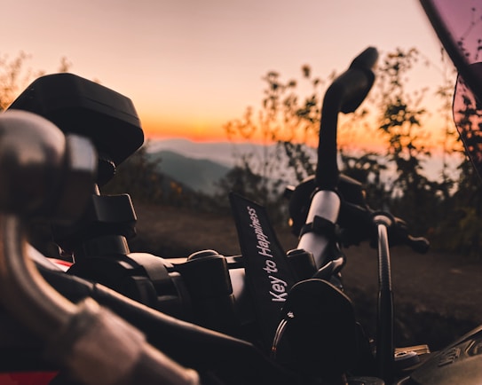 A close-up shot of a motorcycle handlebar with a keychain that reads 'Key to Happiness'. The background features a scenic view of hills and a vibrant sunset, with the sky displaying hues of orange and yellow.
