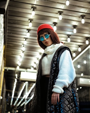 A person stands confidently under a ceiling of bright lights, wearing reflective sunglasses, a red beanie, a thick white sweater, and a long black quilted coat. The setting suggests a modern, possibly urban area.