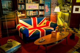 A cozy room with a Union Jack themed sofa surrounded by bookshelves and a wall with a brick pattern. The room features a vintage style with a wooden coffee table, potted plants, and dim lighting that creates a warm atmosphere. A Beefeater London sign sits above the sofa, and the floor is covered with contrasting checkered and striped patterns.