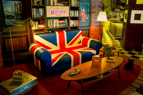 A cozy room with a Union Jack themed sofa surrounded by bookshelves and a wall with a brick pattern. The room features a vintage style with a wooden coffee table, potted plants, and dim lighting that creates a warm atmosphere. A Beefeater London sign sits above the sofa, and the floor is covered with contrasting checkered and striped patterns.