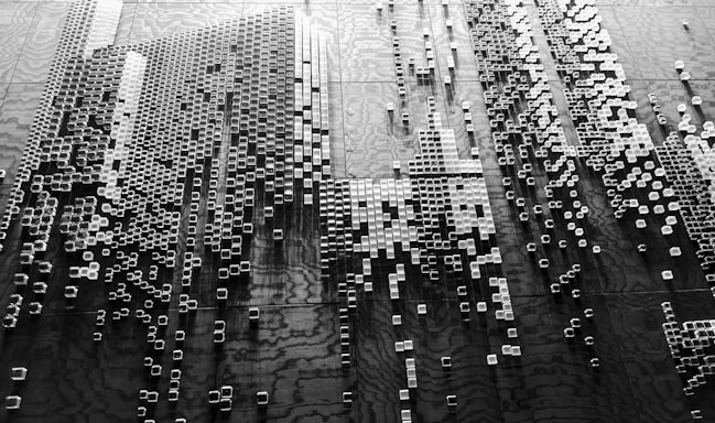 A black and white abstract arrangement of small, three-dimensional square blocks is positioned against a textured background. The blocks vary in height, creating a sense of depth and complexity, with shadows adding to the visual texture. The pattern suggests a digital or futuristic theme, reminiscent of cityscapes or data visualizations.