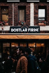 A brick building features a sign indicating 'Dostlar Firini', with an orange awning beneath it displaying the text 'Boyozun ilk adresi'. People are walking by and standing around a bakery or cafe. The facade includes flower boxes with red flowers in the windows.