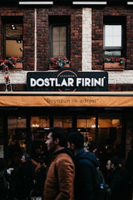 A brick building features a sign indicating 'Dostlar Firini', with an orange awning beneath it displaying the text 'Boyozun ilk adresi'. People are walking by and standing around a bakery or cafe. The facade includes flower boxes with red flowers in the windows.