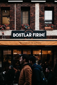 A brick building features a sign indicating 'Dostlar Firini', with an orange awning beneath it displaying the text 'Boyozun ilk adresi'. People are walking by and standing around a bakery or cafe. The facade includes flower boxes with red flowers in the windows.