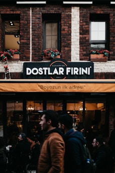 A brick building features a sign indicating 'Dostlar Firini', with an orange awning beneath it displaying the text 'Boyozun ilk adresi'. People are walking by and standing around a bakery or cafe. The facade includes flower boxes with red flowers in the windows.