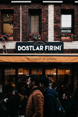 A brick building features a sign indicating 'Dostlar Firini', with an orange awning beneath it displaying the text 'Boyozun ilk adresi'. People are walking by and standing around a bakery or cafe. The facade includes flower boxes with red flowers in the windows.