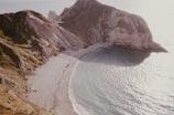 A coastal landscape featuring a secluded beach surrounded by steep cliffs. The beach is covered with pebbles and the sea is calm, reflecting sunlight. The cliffs show distinct layers of rock formations, and patches of grass are visible on the slopes.