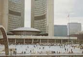 A large public square in front of a significant architectural building with modern curved structures and tall towers. Numerous people are scattered across the open space, engaged in leisure activities. A flag on a tall pole is visible, and surrounding office buildings highlight an urban environment.
