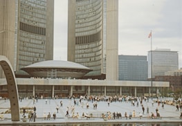 A large public square in front of a significant architectural building with modern curved structures and tall towers. Numerous people are scattered across the open space, engaged in leisure activities. A flag on a tall pole is visible, and surrounding office buildings highlight an urban environment.