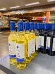 A display of wine bottles is neatly arranged on a store shelf. There are two types of wine visible: white wine with yellow liquid and blue caps, and red wine with dark bottles and blue caps. The background shows a modern wine shop interior with wooden accents and the word 'adega' written above.