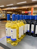 A display of wine bottles is neatly arranged on a store shelf. There are two types of wine visible: white wine with yellow liquid and blue caps, and red wine with dark bottles and blue caps. The background shows a modern wine shop interior with wooden accents and the word 'adega' written above.
