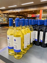 A display of wine bottles is neatly arranged on a store shelf. There are two types of wine visible: white wine with yellow liquid and blue caps, and red wine with dark bottles and blue caps. The background shows a modern wine shop interior with wooden accents and the word 'adega' written above.