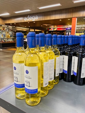 A display of wine bottles is neatly arranged on a store shelf. There are two types of wine visible: white wine with yellow liquid and blue caps, and red wine with dark bottles and blue caps. The background shows a modern wine shop interior with wooden accents and the word 'adega' written above.