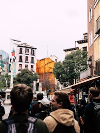 A bustling street scene with a group of people walking towards a square. The buildings are adorned with colorful murals, including one featuring figures holding a plant, and a striking orange facade. Trees and lampposts line the street, creating a lively urban environment.