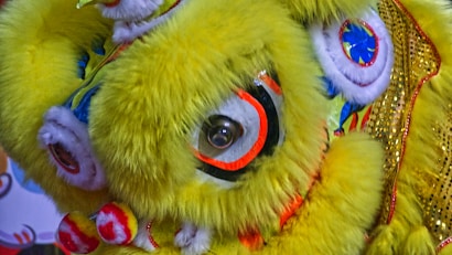 A bright and colorful lion dance costume with vibrant yellow fur and detailed facial features. The costume is adorned with striking eyes and intricate patterns in red, blue, and white. It is embellished with shimmering gold sequins.