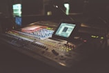 A dimly lit audio mixing console with numerous buttons and knobs, featuring a digital screen displaying software for sound editing. The equipment occupies a central position and is surrounded by a warm, ambient glow.
