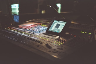 A dimly lit audio mixing console with numerous buttons and knobs, featuring a digital screen displaying software for sound editing. The equipment occupies a central position and is surrounded by a warm, ambient glow.