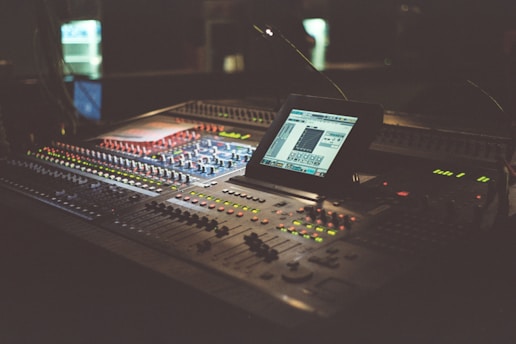 A dimly lit audio mixing console with numerous buttons and knobs, featuring a digital screen displaying software for sound editing. The equipment occupies a central position and is surrounded by a warm, ambient glow.