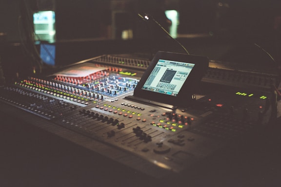 A dimly lit audio mixing console with numerous buttons and knobs, featuring a digital screen displaying software for sound editing. The equipment occupies a central position and is surrounded by a warm, ambient glow.