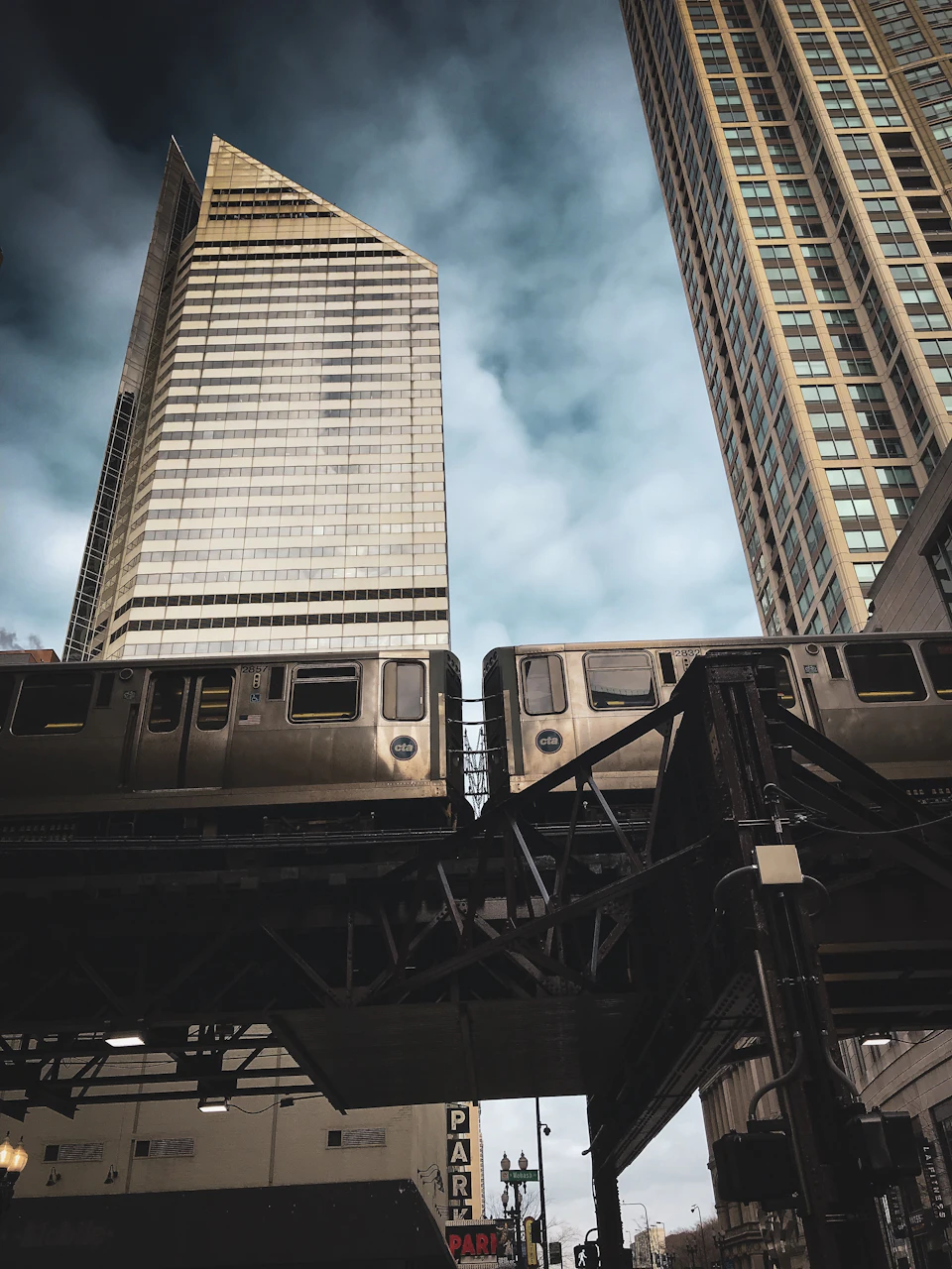 Two trains cross paths over elevated metal frame in cityscape with tall buildings nearby