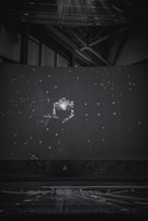 A dark setting with a large spherical object surrounded by numerous small, bright dots resembling stars. The scene resembles a projection or display, possibly in a planetarium or a dome. Industrial structures and beams can be seen faintly in the background.