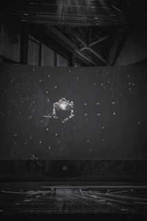 A dark setting with a large spherical object surrounded by numerous small, bright dots resembling stars. The scene resembles a projection or display, possibly in a planetarium or a dome. Industrial structures and beams can be seen faintly in the background.