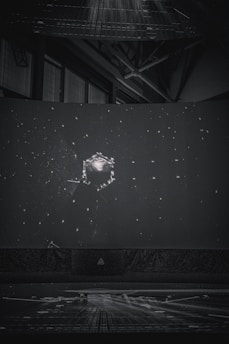 A dark setting with a large spherical object surrounded by numerous small, bright dots resembling stars. The scene resembles a projection or display, possibly in a planetarium or a dome. Industrial structures and beams can be seen faintly in the background.