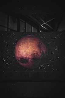 A planet with a reddish hue and detailed craters is set against a backdrop of stars in a dark, cosmic environment. The image contains an intriguing play of light and shadow on the planet's surface, highlighting its texture.