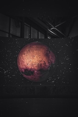 A planet with a reddish hue and detailed craters is set against a backdrop of stars in a dark, cosmic environment. The image contains an intriguing play of light and shadow on the planet's surface, highlighting its texture.
