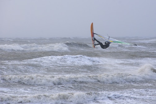 A windsurfer is captured in mid-air above choppy ocean waves under a cloudy sky. The person is wearing a wetsuit and holding onto a brightly colored sail. The sea is tumultuous, and the sky appears overcast, adding a dramatic effect to the scene.