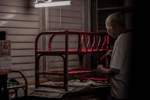 An elderly person is painting a red wooden structure under a harsh white ceiling light, surrounded by a dimly lit, wooden interior. Newspapers cover the surface beneath the structure.