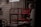 An elderly person is painting a red wooden structure under a harsh white ceiling light, surrounded by a dimly lit, wooden interior. Newspapers cover the surface beneath the structure.