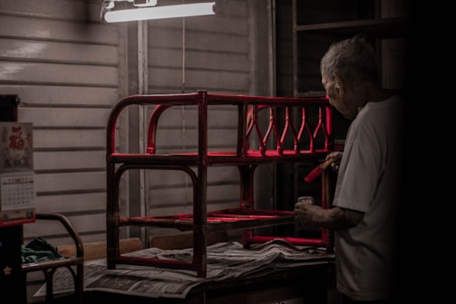 An elderly person is painting a red wooden structure under a harsh white ceiling light, surrounded by a dimly lit, wooden interior. Newspapers cover the surface beneath the structure.